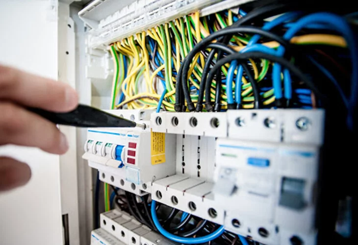 Mão masculina segurando uma ferramenta de manutenção em um quadro de luz aberto com os fios coloridos a mostra.